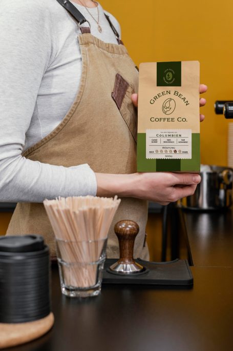 Barista hält eine Packung Kaffee in der Hand, neben einem Glas mit Holzstäbchen.